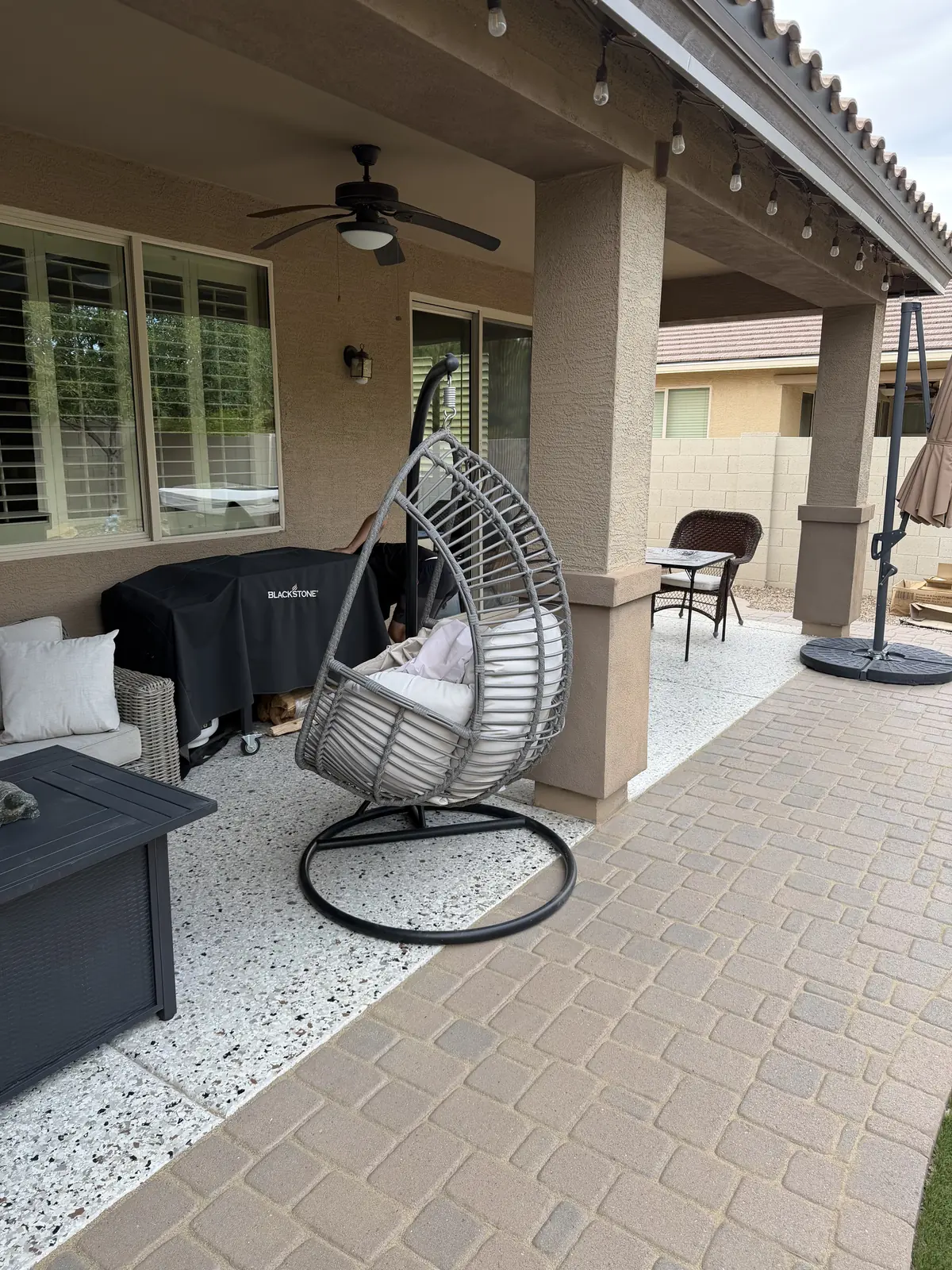 A covered patio with a hanging egg chair, a grill and string lights above clean pavers